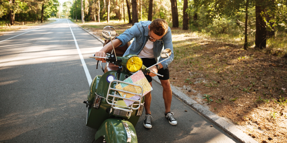 Un hombre de pie junto a una motocicleta verde en una carretera rodeada de árboles. El hombre está observando un mapa que tiene sujeto al manillar de la motocicleta. La carretera está vacía y se extiende en línea recta hacia el horizonte.