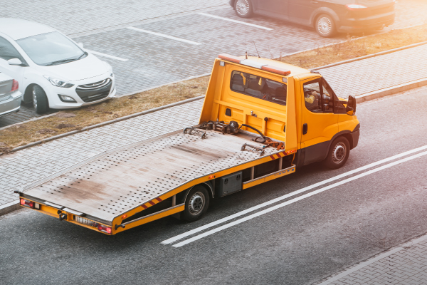 Un camión de remolque amarillo está estacionado en una carretera con dos líneas blancas continuas. El camión tiene una plataforma plana y vacía, diseñada para transportar vehículos. En el fondo, se pueden ver varios coches estacionados en un aparcamiento pavimentado. La imagen muestra un entorno urbano con luz solar suave.