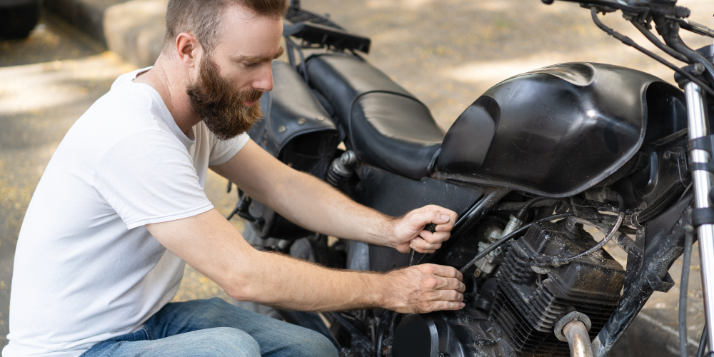 Un hombre con una camiseta blanca y jeans está reparando una motocicleta negra. Utiliza una herramienta para ajustar una parte del motor. La imagen se centra en la actividad de reparación, destacando la interacción del hombre con la motocicleta.