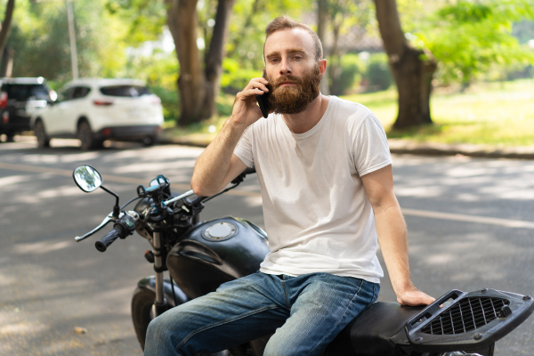 Un hombre con barba y camiseta blanca está sentado en una motocicleta negra mientras habla por teléfono. La imagen fue tomada en una calle con árboles y coches estacionados al fondo.