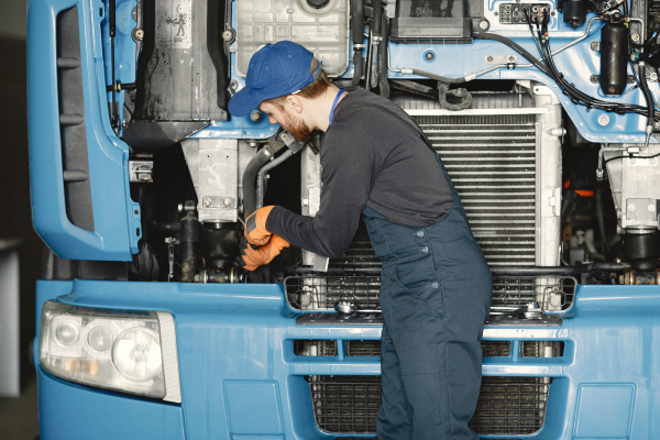 Un mecánico con guantes naranjas y una gorra azul está trabajando en el motor de un camión azul. El capó del camión está levantado, mostrando el radiador y otras partes del motor. El mecánico está inclinado hacia adelante, aparentemente ajustando o reparando una parte del motor.