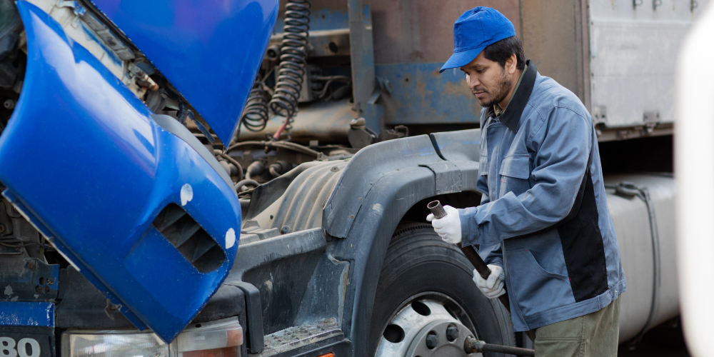 Un hombre con una gorra azul y una chaqueta de trabajo está revisando o reparando un camión. El capó del camión, que es de color azul, está levantado, y el hombre sostiene una herramienta en la mano derecha mientras se inclina hacia la rueda delantera del camión. La imagen muestra una escena de mantenimiento o reparación de vehículos, lo cual es relevante para ilustrar trabajos de mecánica o mantenimiento de camiones.