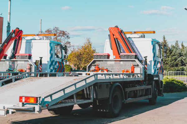 Dos camiones grúa con rampas inclinadas en un área abierta. Ambos camiones tienen brazos hidráulicos de color naranja y están equipados con luces de advertencia en la parte superior. Las rampas están diseñadas para cargar y descargar vehículos. En el fondo, se pueden ver árboles y un cielo despejado. Esta imagen es relevante porque muestra vehículos especializados utilizados para el transporte y remolque de otros vehículos, lo cual es esencial en situaciones de emergencia o para el transporte de vehículos averiados.