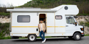 La imagen muestra una autocaravana blanca estacionada en una carretera con un paisaje verde y montañoso de fondo. La autocaravana tiene una franja amarilla horizontal a lo largo de su costado. La puerta de la autocaravana está abierta y una persona está de pie junto a ella, apoyada en el marco de la puerta. La persona lleva una chaqueta beige, una camiseta blanca, jeans azules y zapatillas blancas.