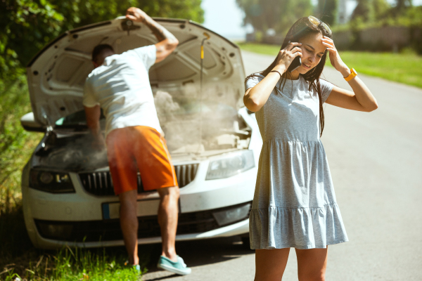 Dos personas junto a un automóvil averiado en una carretera. Una persona está revisando el motor del coche, que tiene el capó levantado y parece estar echando humo. La otra persona está de pie cerca del coche, con una mano en la cabeza y la otra sosteniendo un teléfono móvil, posiblemente llamando para pedir ayuda. La escena es interesante porque muestra una situación común de emergencia en la carretera, donde un coche ha sufrido una avería y las personas están tratando de resolver el problema.